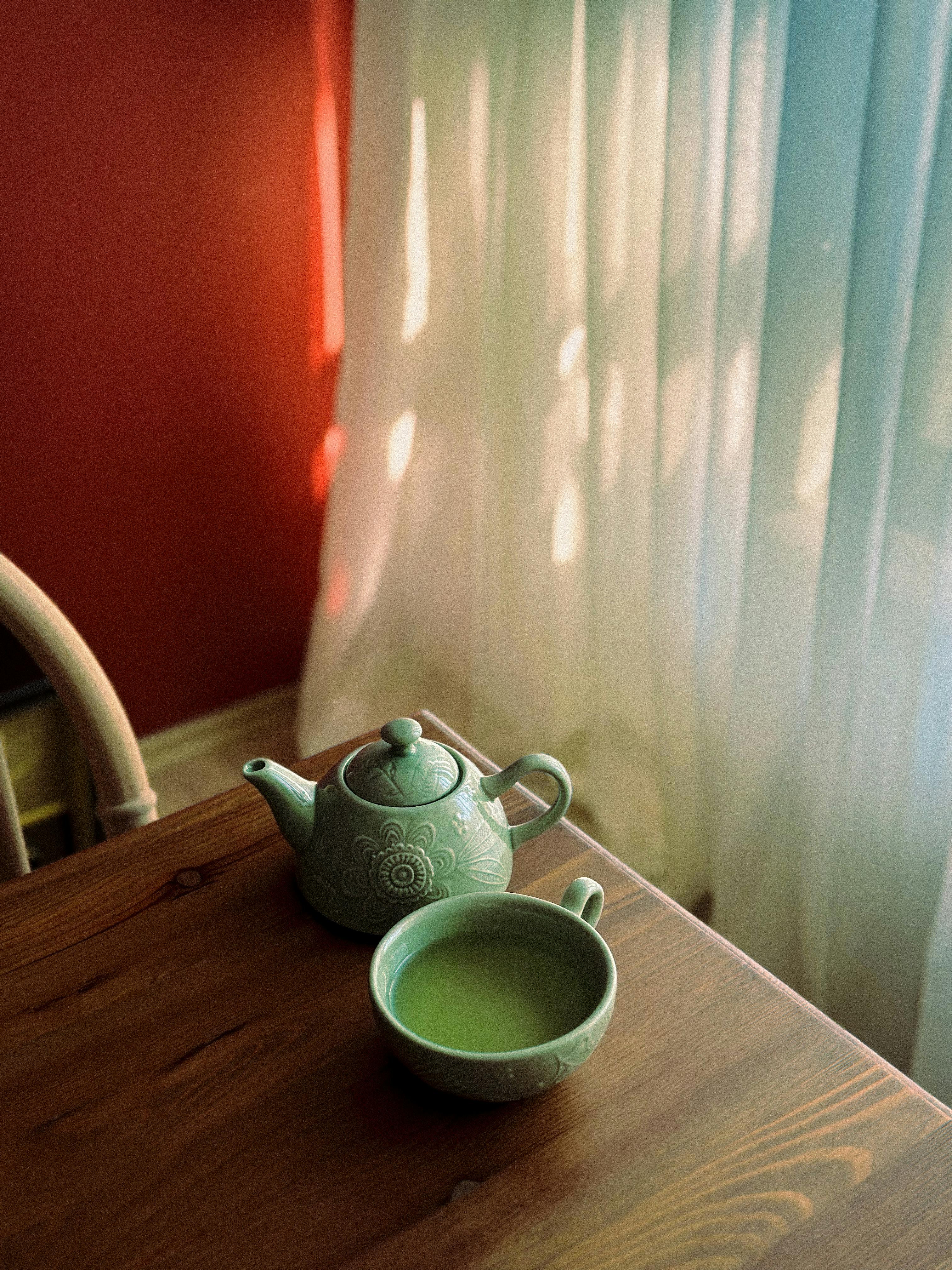 Green ceramic teapot and cup with green tea on a wooden table by a window with sheer curtains.