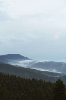 A serene view of misty forested mountains with clouds under a cloudy sky.