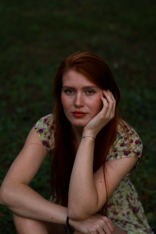 Free Close-up portrait of a redhead woman sitting outdoors in a floral dress, looking directly at the camera. Stock Photo