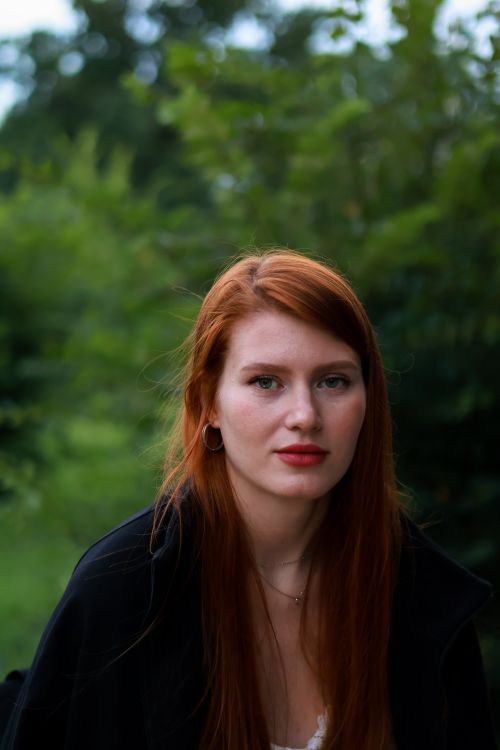 Free A young redhead woman with red lips poses outdoors surrounded by lush greenery. Stock Photo