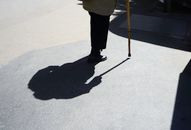 Silhouette of senior walking with a cane on sunlit pavement, highlighting shadows.