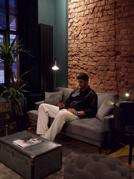 Adult man sitting on a sofa using a smartphone in a modern loft with brick wall and indoor plants.