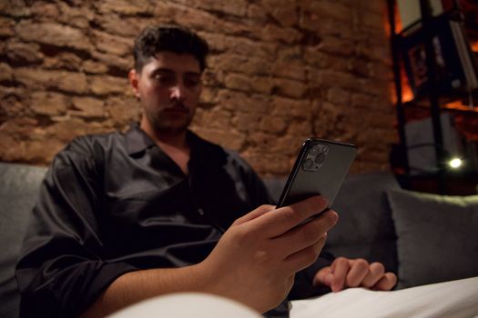 Young man relaxing on a couch while using a smartphone at night indoors.