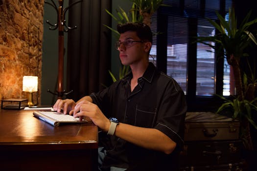 Adult male wearing eyeglasses typing on keyboard in a dimly lit room with plants.