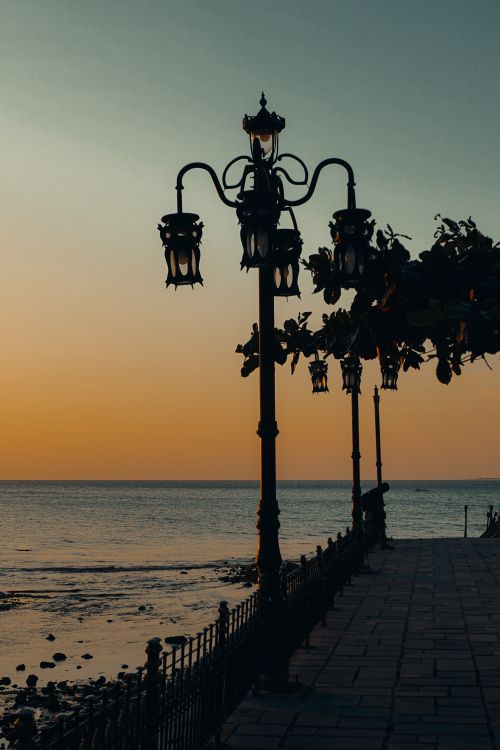 Free Captivating sunset by the sea with ornate street lamps casting silhouettes for a tranquil scene. Stock Photo