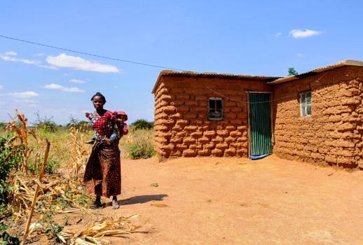 A mother and child outside a traditional mud house in Ihumwa, Dodoma, Tanzania, showcasing rural life.