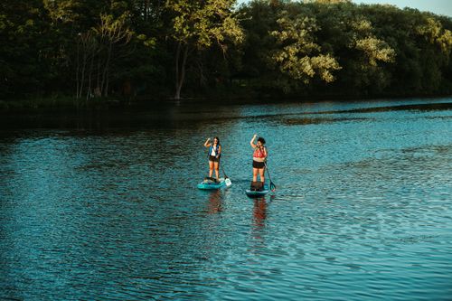Free Two women paddleboarding on a serene lake surrounded by lush greenery under a clear sky. Stock Photo