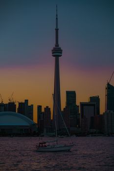 Stunning silhouette of Toronto skyline at sunset with iconic CN Tower and sailboat on calm water.