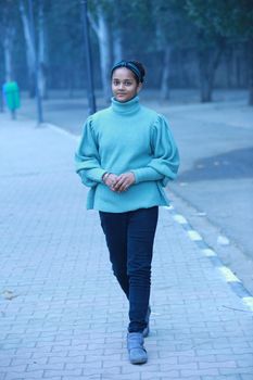 A young woman walks along a tree-lined street in Chandigarh during fall, wearing a stylish sweater.