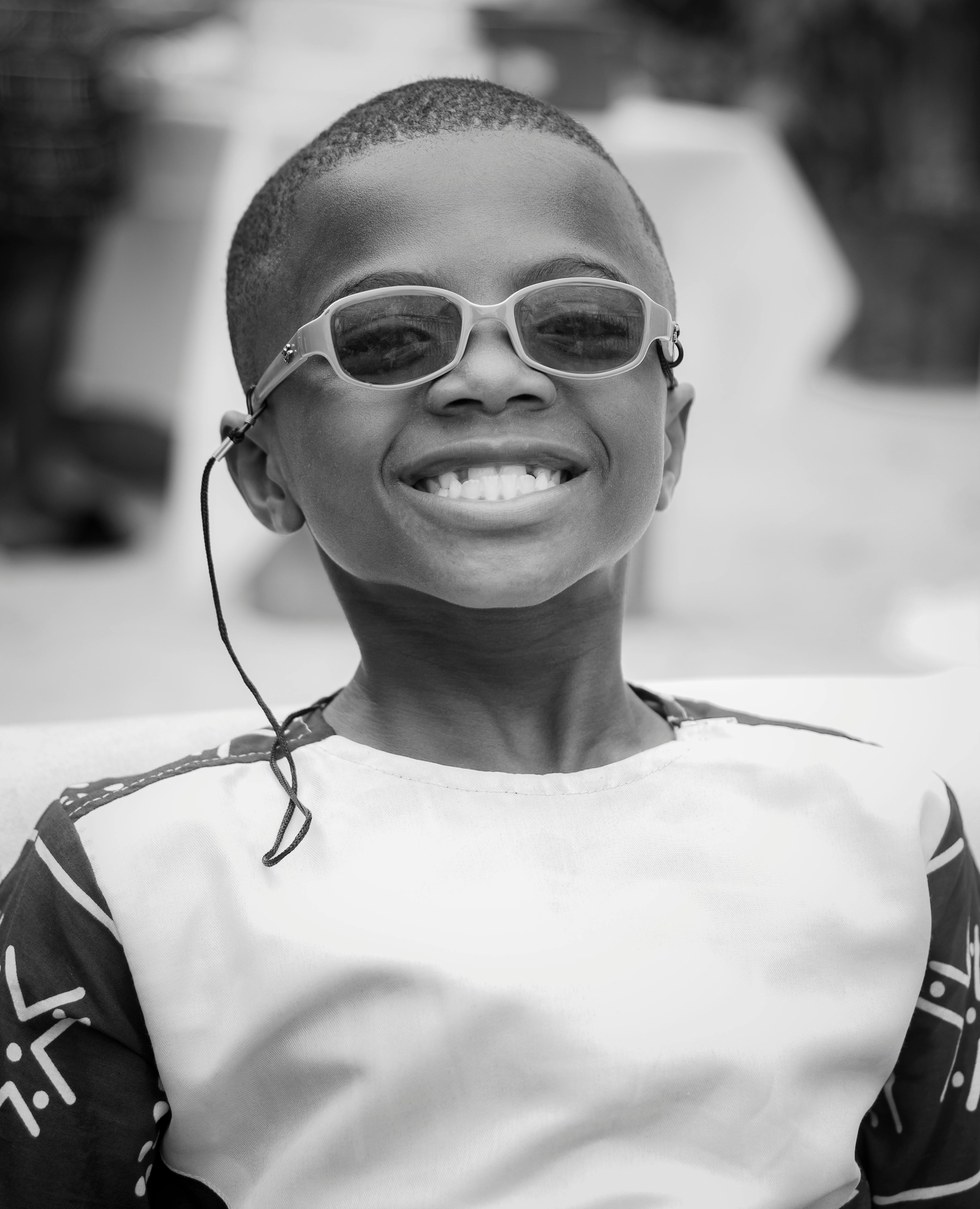 A cheerful boy wearing sunglasses smiles brightly in an outdoor portrait.
