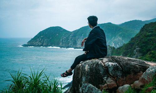 Free A man sits on a clifftop, gazing at the serene ocean and lush green mountains. Stock Photo