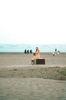 A woman in a yellow dress and hat sits alone on a beach with a serene sea in the background.