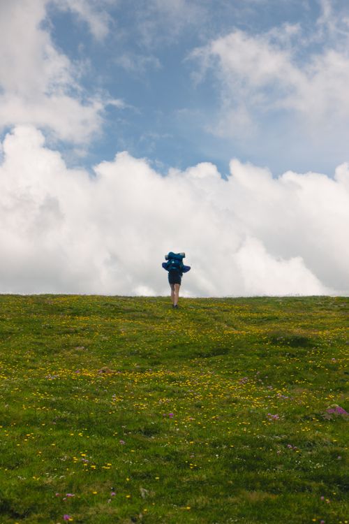 Free Lone hiker with backpack walking uphill through a grassy field under a dramatic sky. Stock Photo
