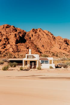 A solitary house stands amidst the arid desert sands and red rocky formations under a clear blue sky.