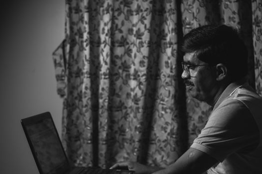 A black and white portrait of a man using a laptop indoors, focusing intently.