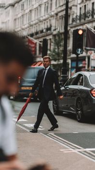Stylish businessman with umbrella crossing city street in London, UK. Urban lifestyle concept.
