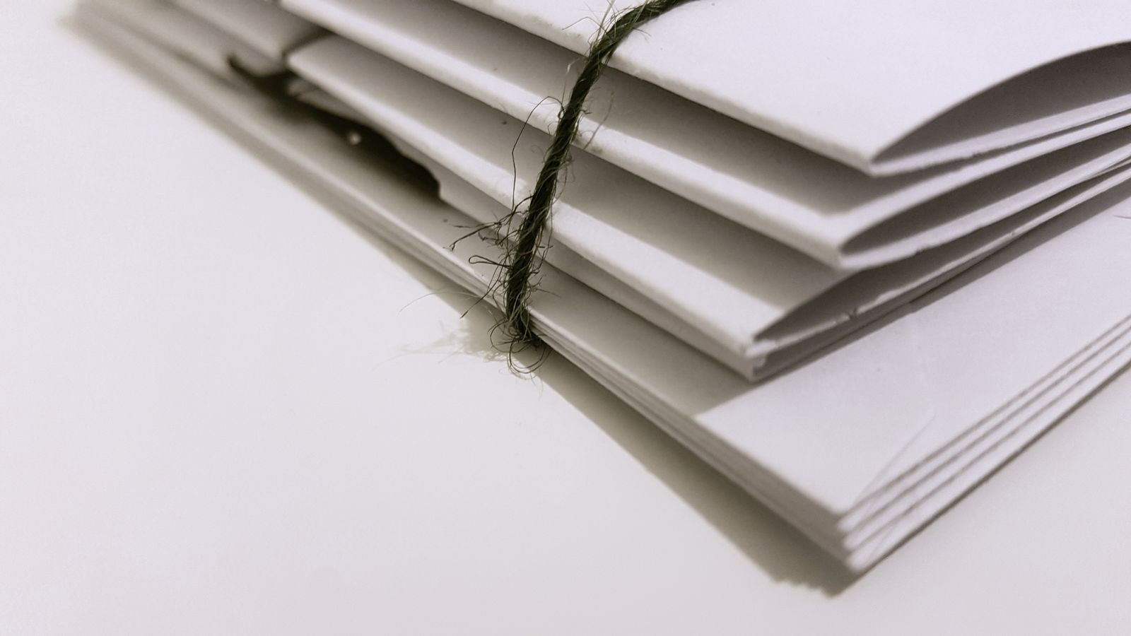 A neat stack of plain white envelopes on a white surface, bound in the middle by a length of dark green twine tied in a knot