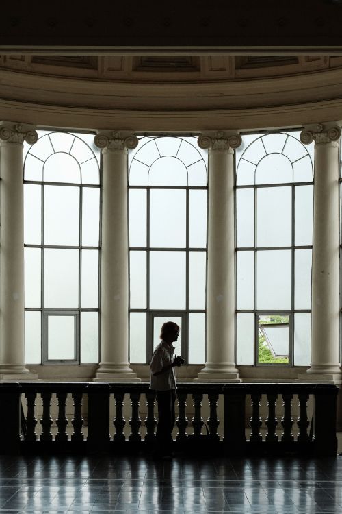 Free A lone person silhouetted against tall arched windows in a historic Ho Chi Minh City museum. Stock Photo