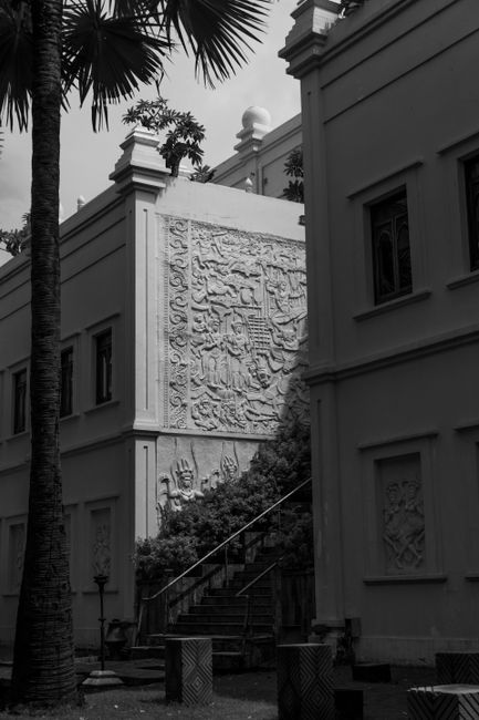 A black and white photo capturing intricate bas relief on city building wall with stairs and palm tree.