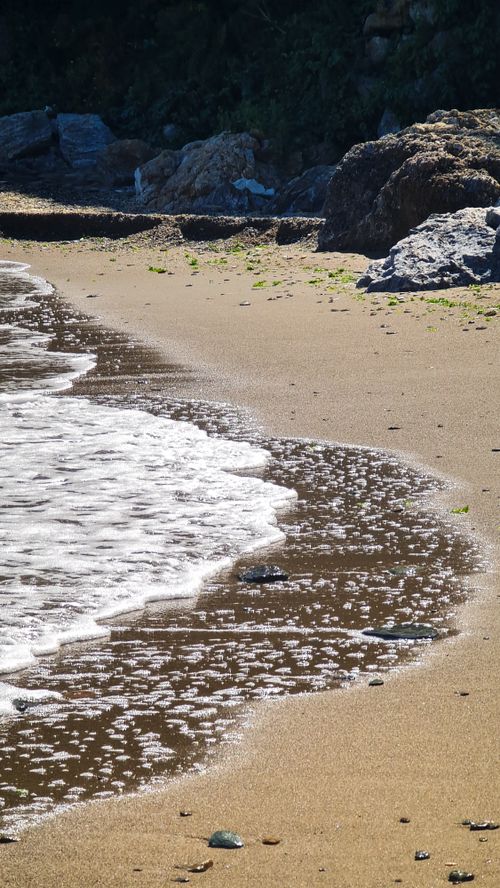 Free A peaceful sandy beach where waves gently meet the shore with rocks in the background. Stock Photo
