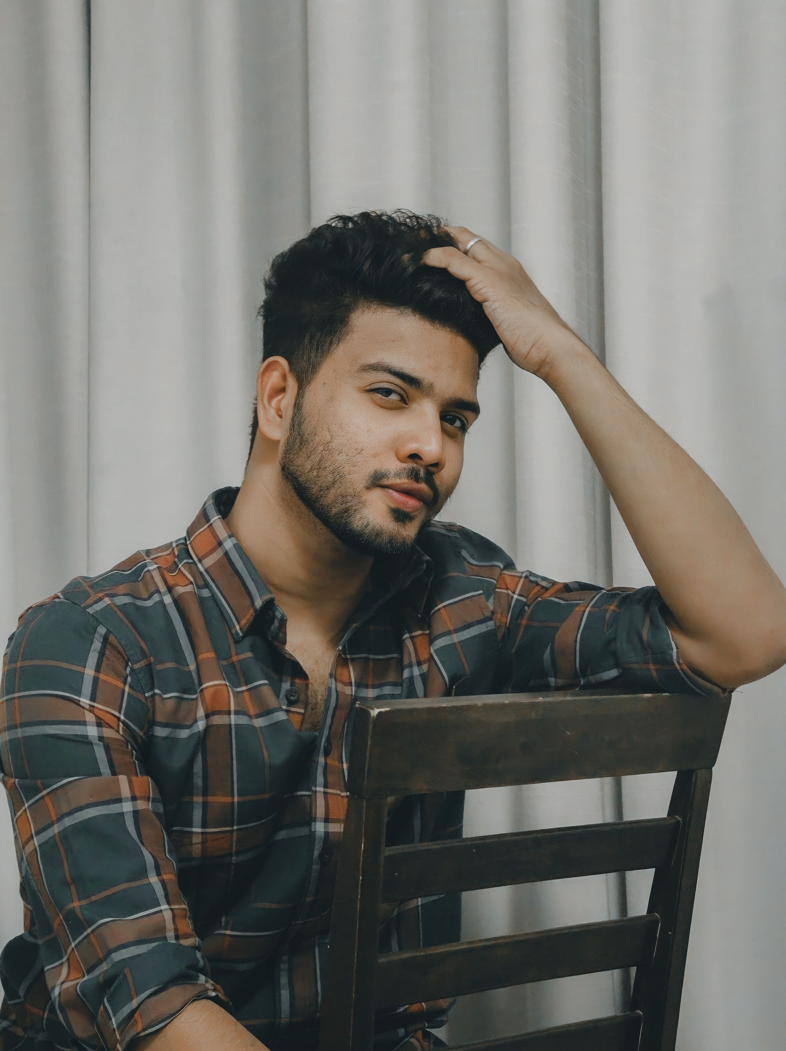 Casual portrait of a man in plaid shirt sitting on a chair with a thoughtful pose indoors.