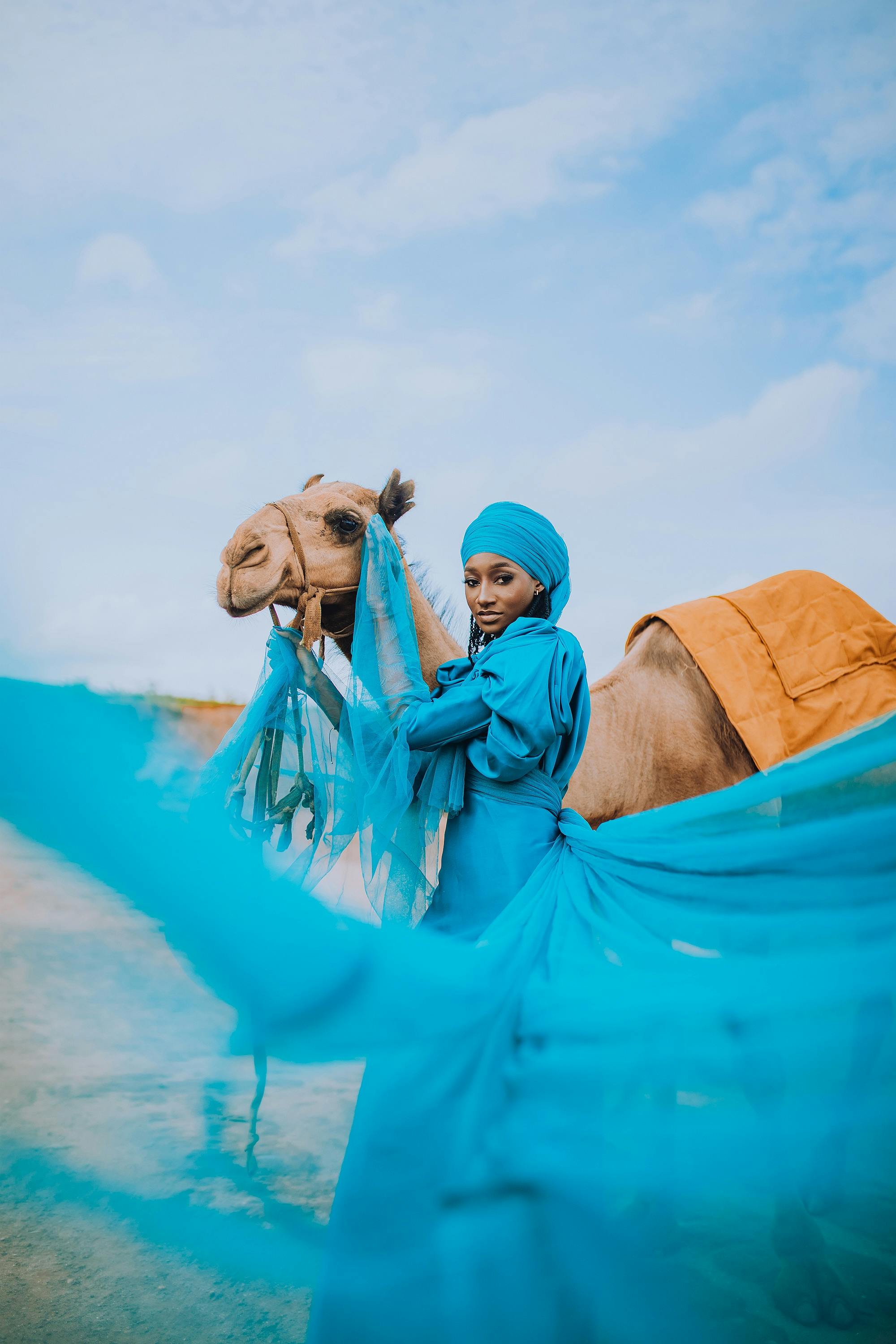 Portrait of a woman in a blue dress with a camel in an outdoor setting.