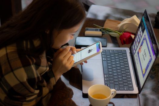 A young woman using a smartphone and laptop at a cozy indoor setting, enjoying a cup of tea.