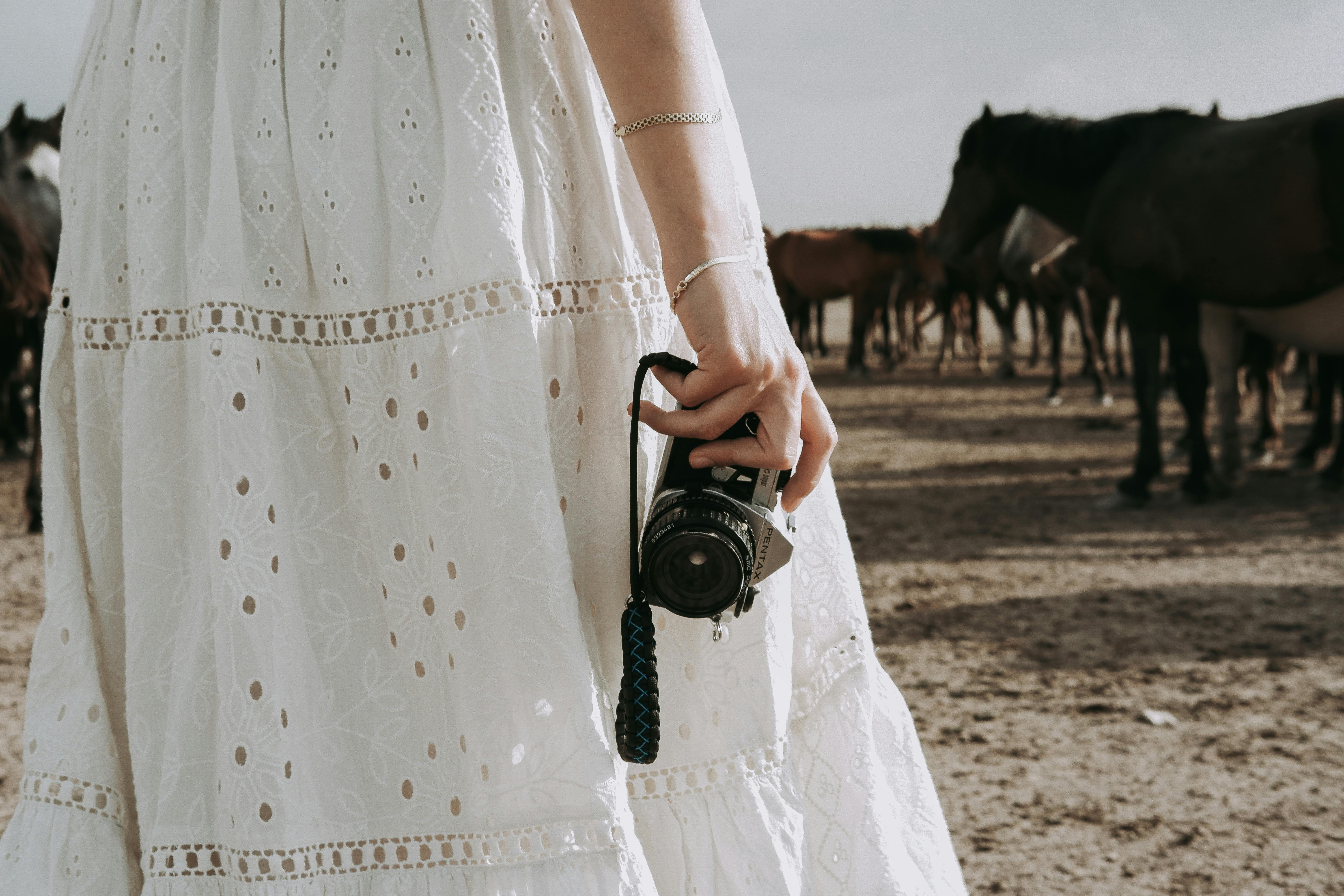 A woman holding a vintage camera stands among a herd of horses outdoors in sunlight.