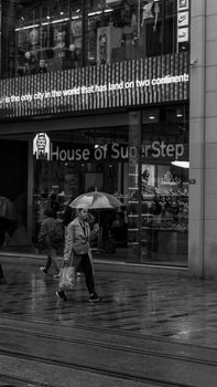 Monochrome image of people walking in rain with umbrellas in urban setting.