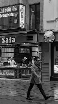 A man walks on a rainy city street in front of local shops, captured in black and white.