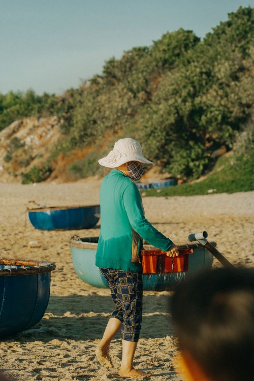 Free A person wearing a hat walks on a sunny beach carrying a box, surrounded by boat baskets. Stock Photo