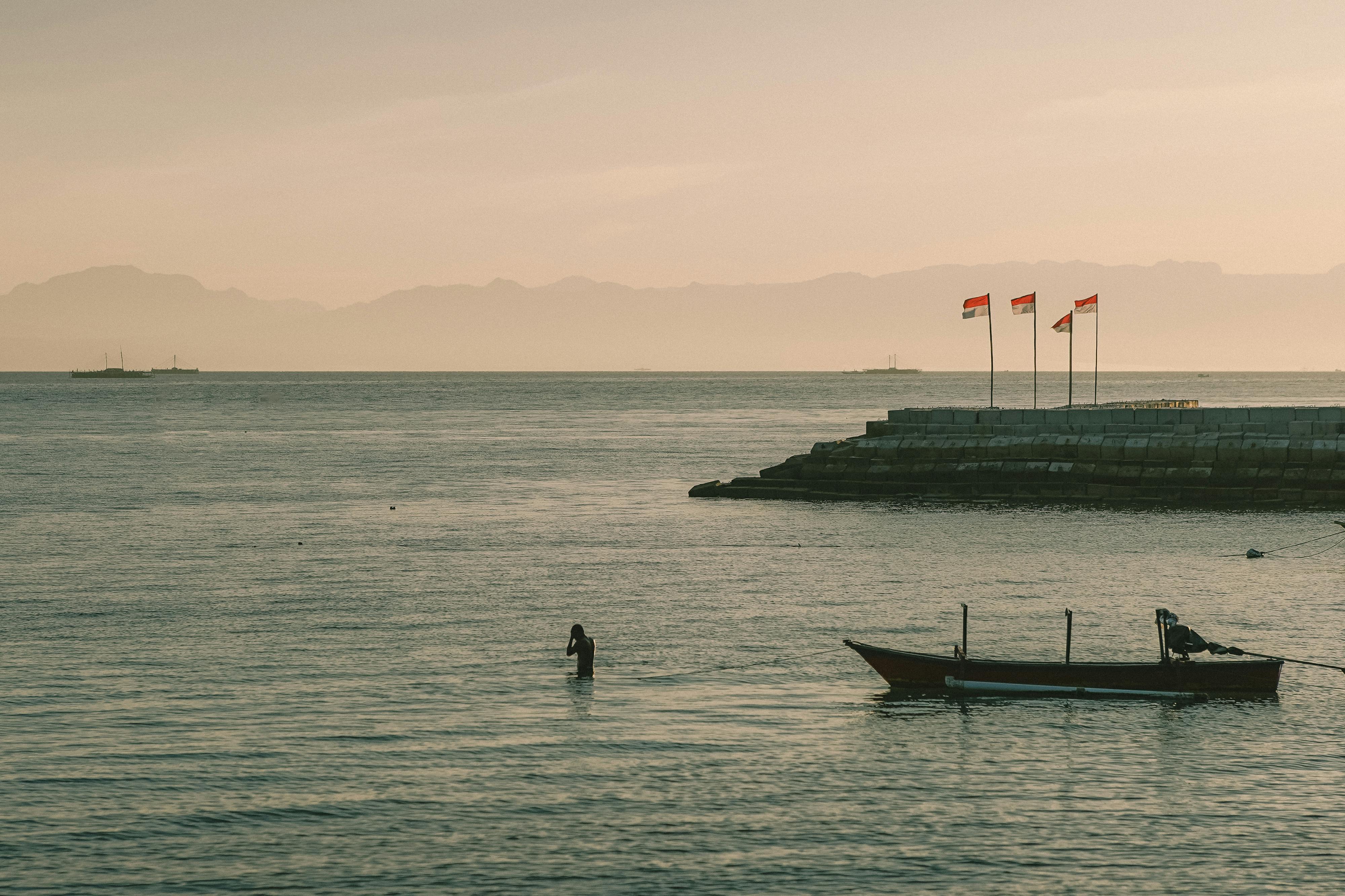 Peaceful evening at Kupang's shoreline with a boat and Indonesian flags, featuring a calm sea view.