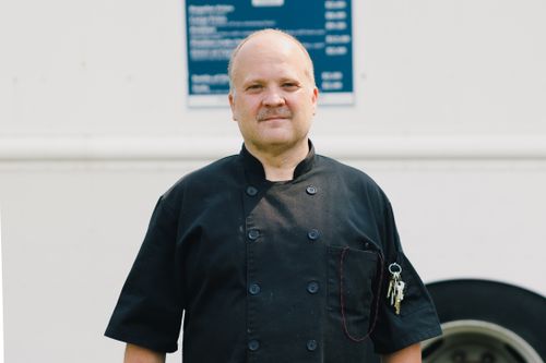 Free Portrait of a professional chef standing confidently in front of a food truck, wearing a black uniform. Stock Photo