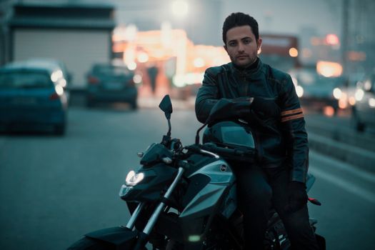 A stylish man poses on a motorcycle in an urban street during twilight.