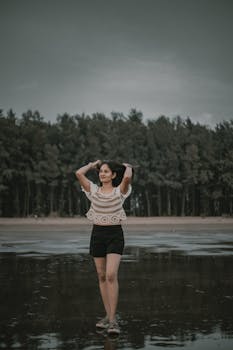 Woman posing on a serene beach with a forest backdrop, embodying calm fashion with an earthy tone.