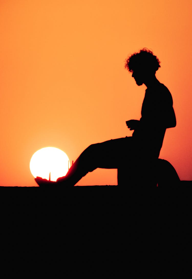 Silhouette Of Man Sitting Near Sun At Sunset On Clear, Yellow Sky