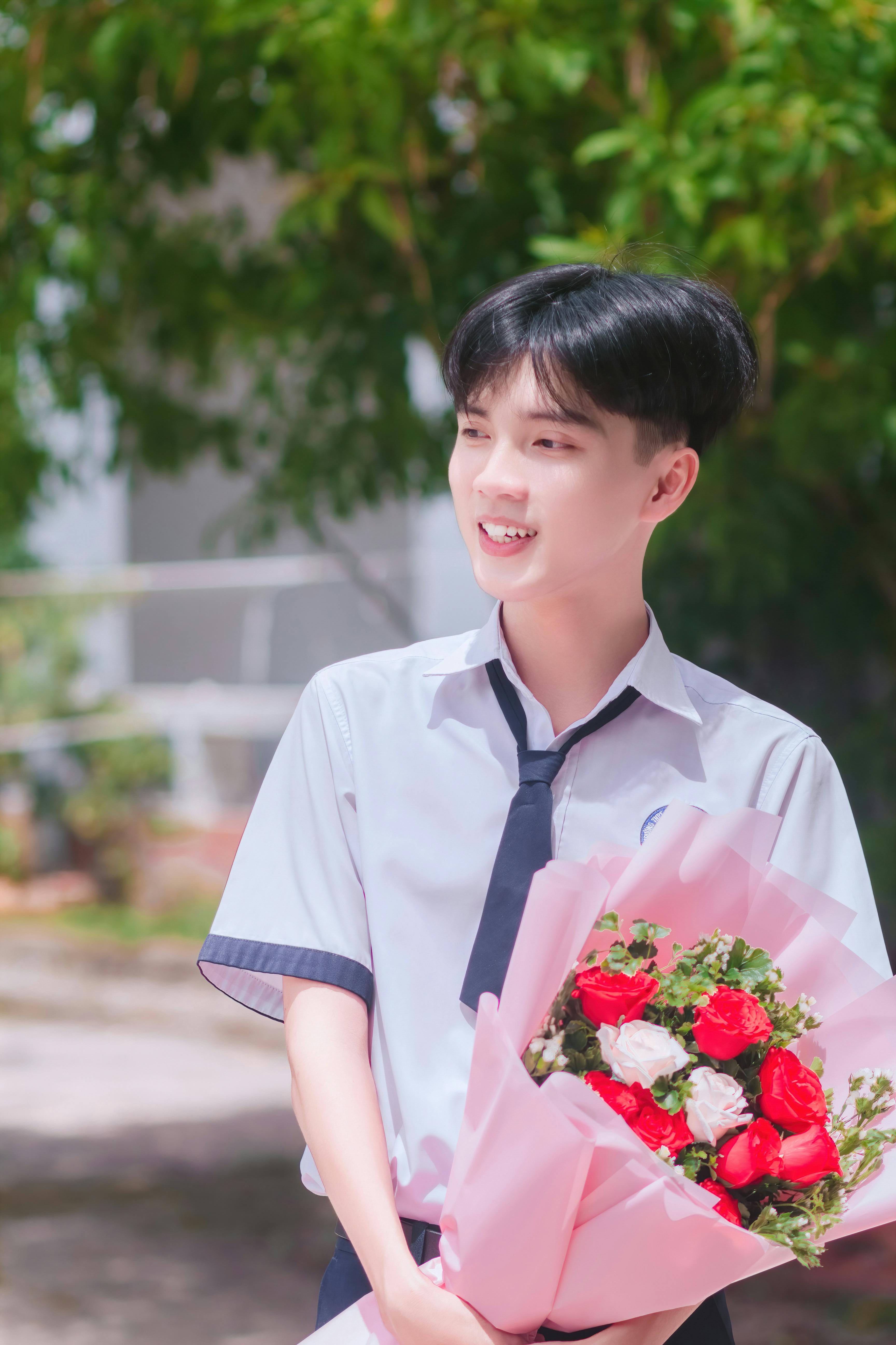 Asian male student in uniform holding a vibrant bouquet, smiling brightly outdoors.