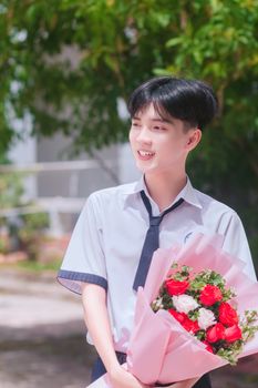 Asian male student in uniform holding a vibrant bouquet, smiling brightly outdoors.