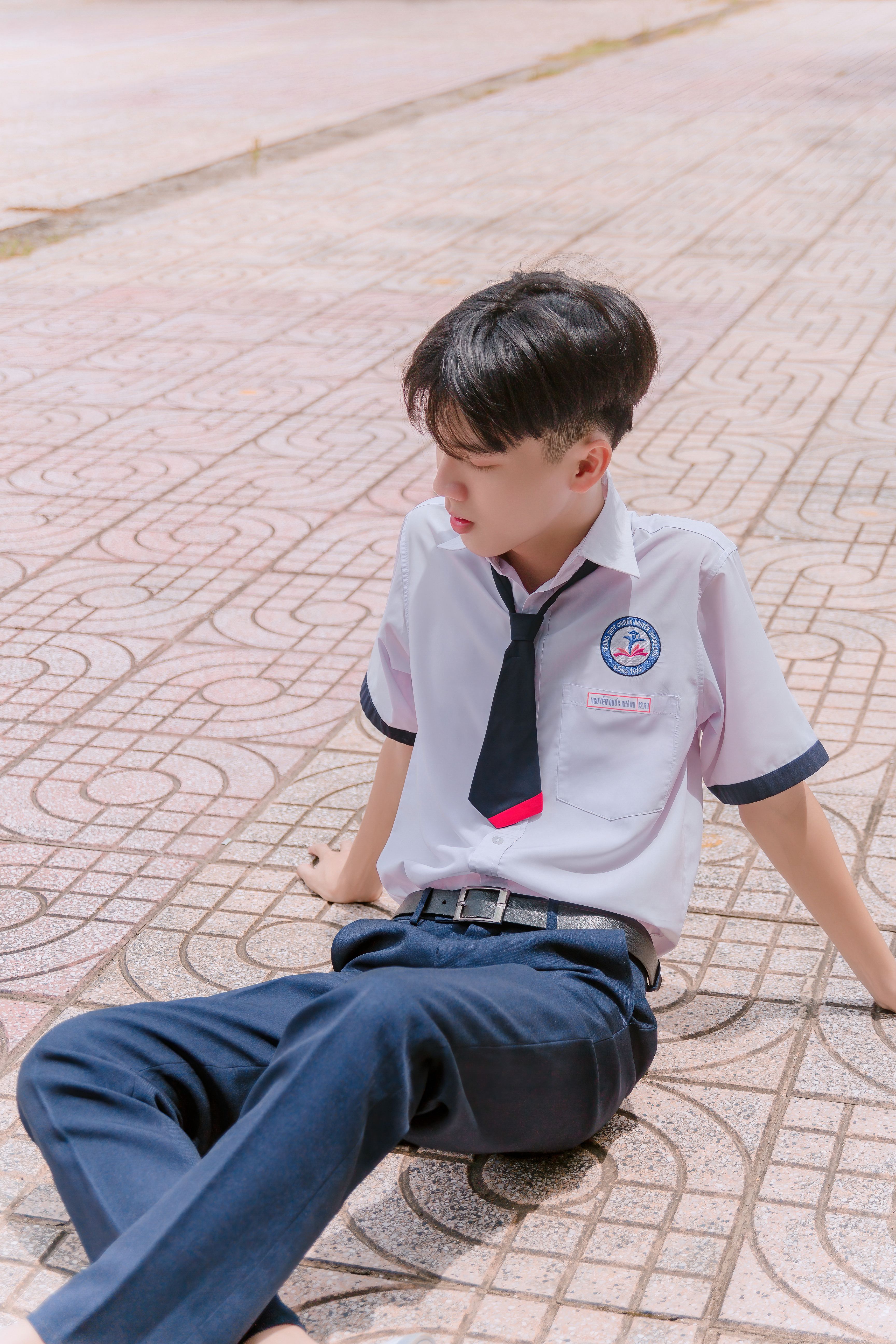 Young man in school uniform sitting on a patterned sidewalk in a relaxed pose.