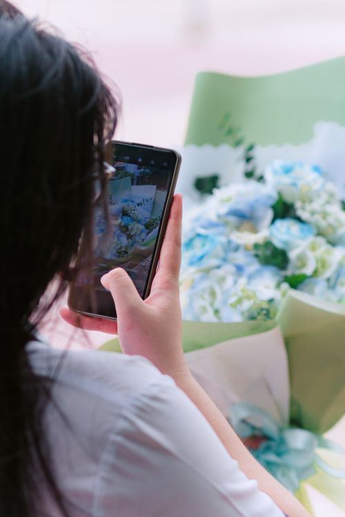 Free A woman captures a close-up photo of a vibrant blue flower bouquet with her smartphone indoors. Stock Photo