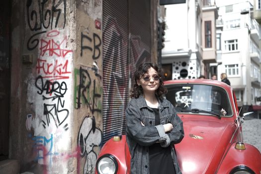 Woman in denim jacket poses by a vintage red Volkswagen Beetle with urban graffiti backdrop.