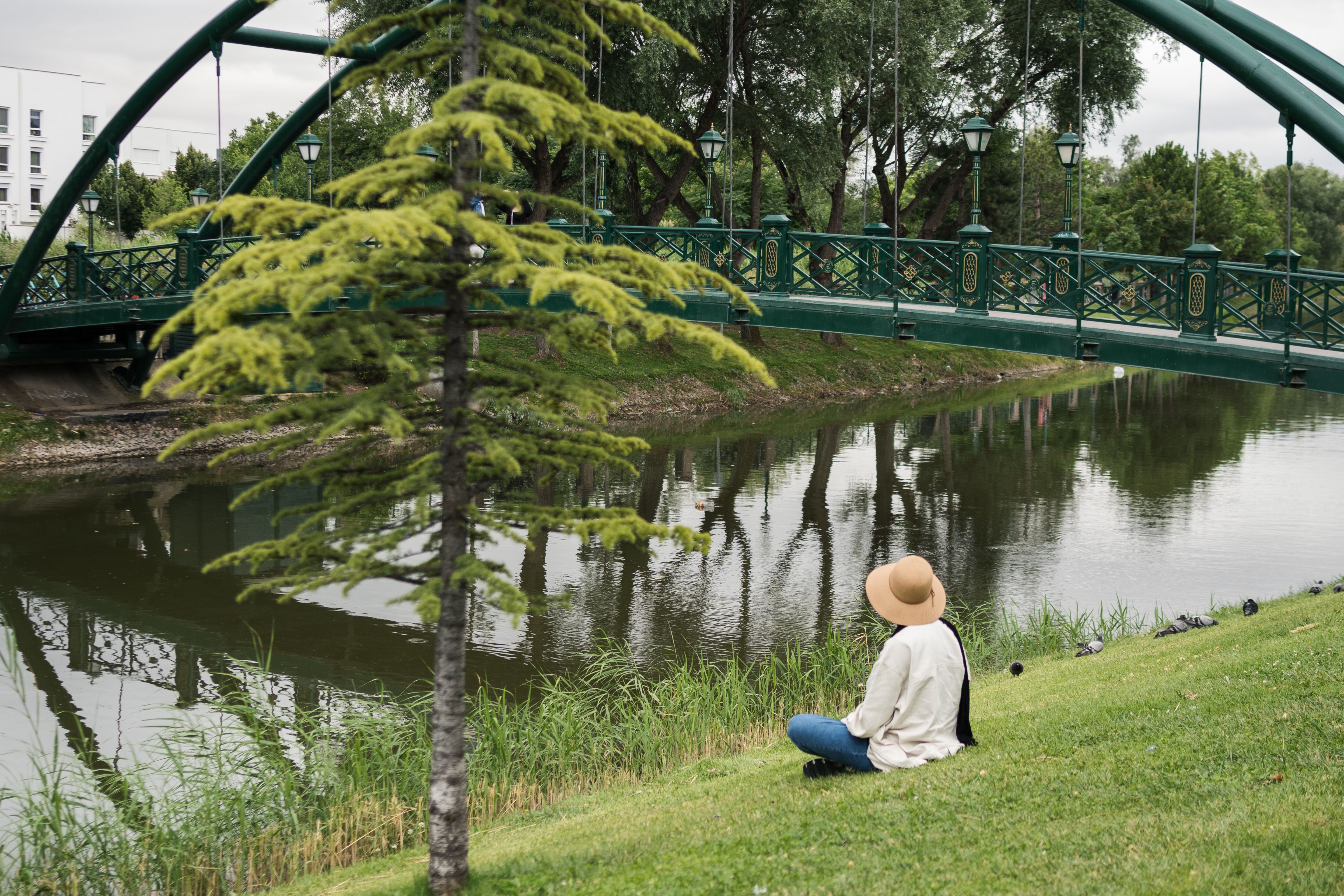 A woman sits by a peaceful creek under a green bridge in Eskişehir, Türkiye.