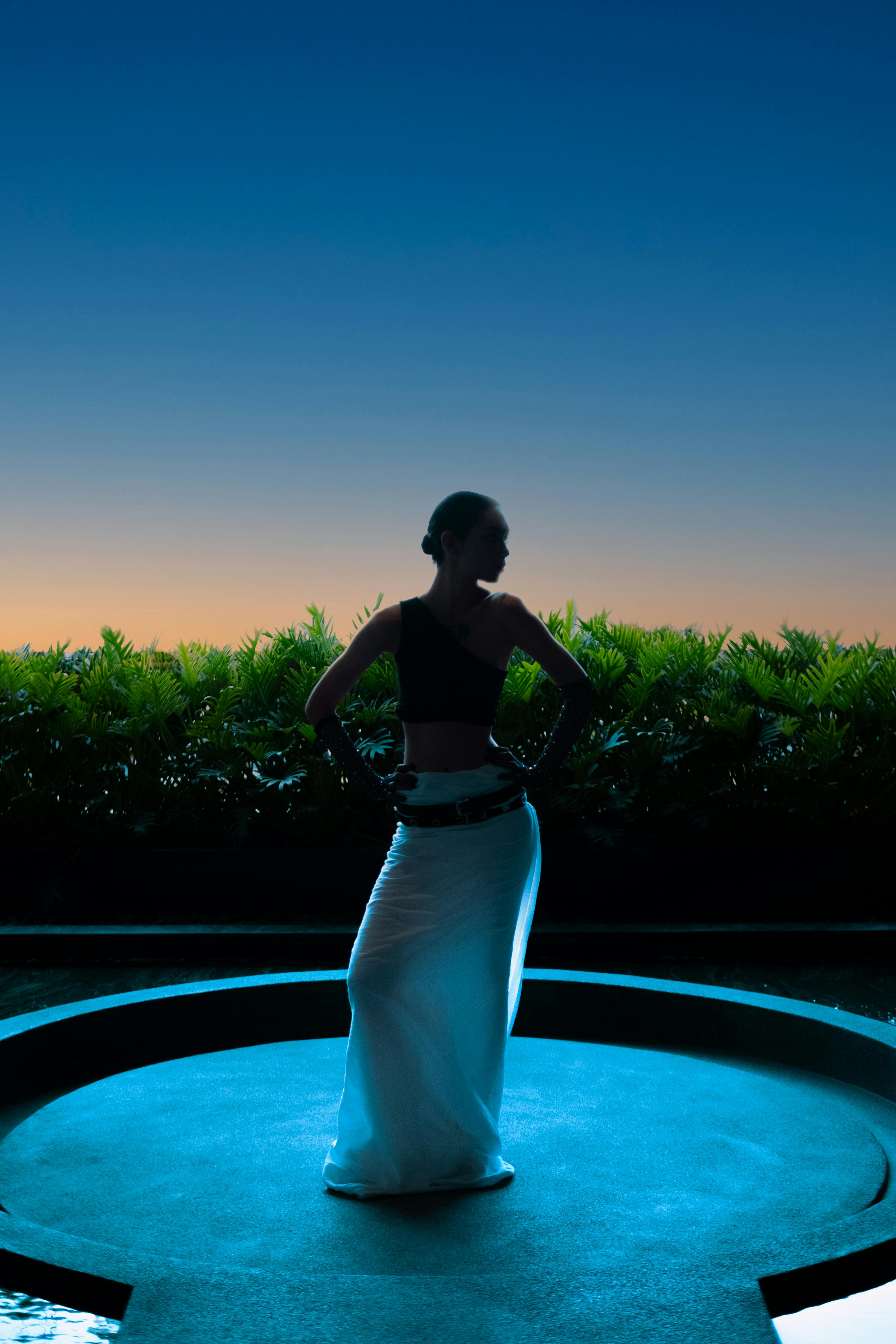 A stylish silhouette of a model in a white skirt standing outdoors at sunset in Bangkok.