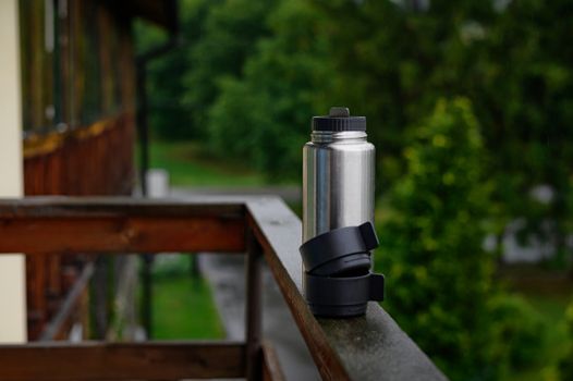 A stainless steel water bottle placed on a wooden balcony railing during a rainy day outdoors.