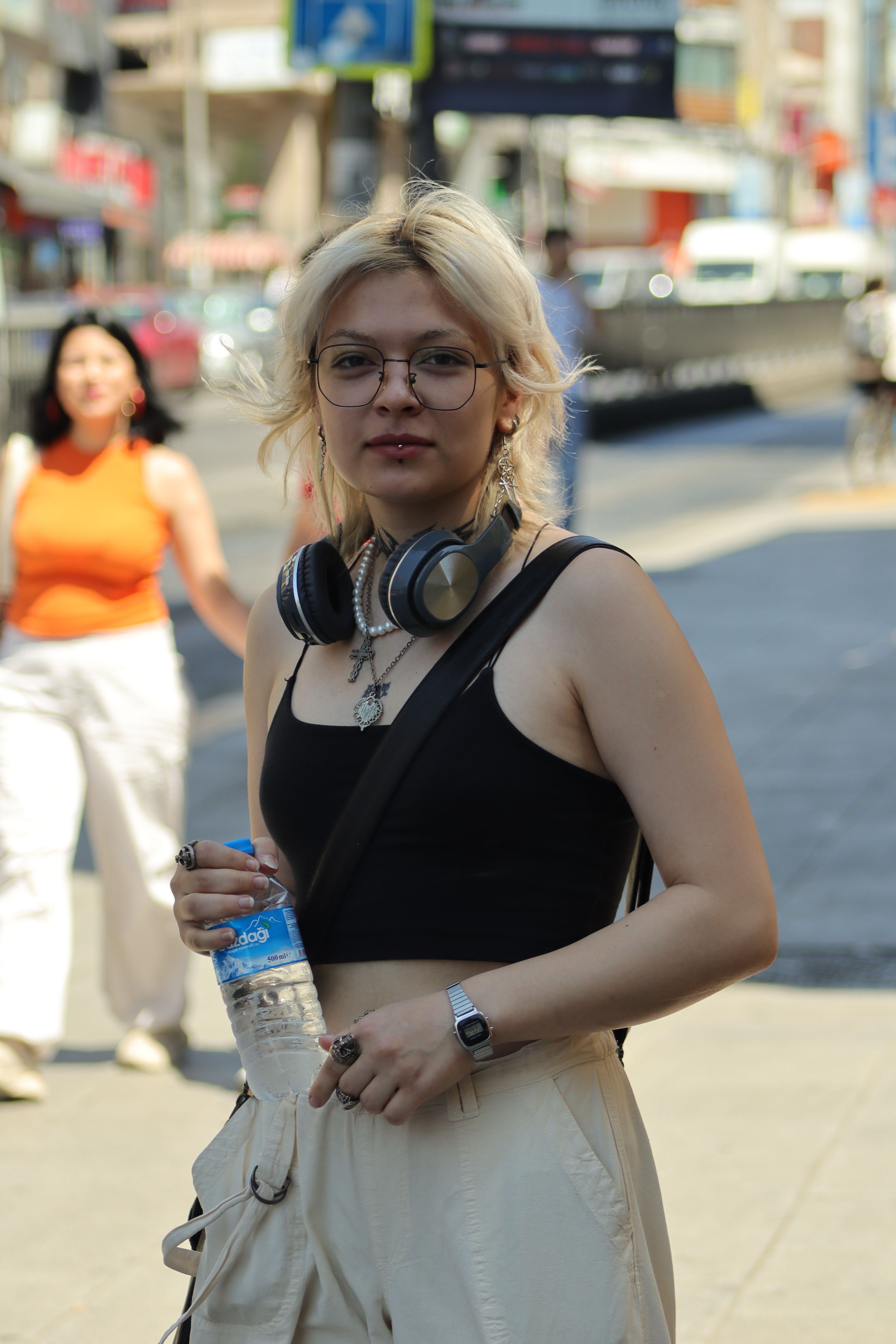 Stylish young woman with headphones and water bottle walking in a busy city street on a sunny day.