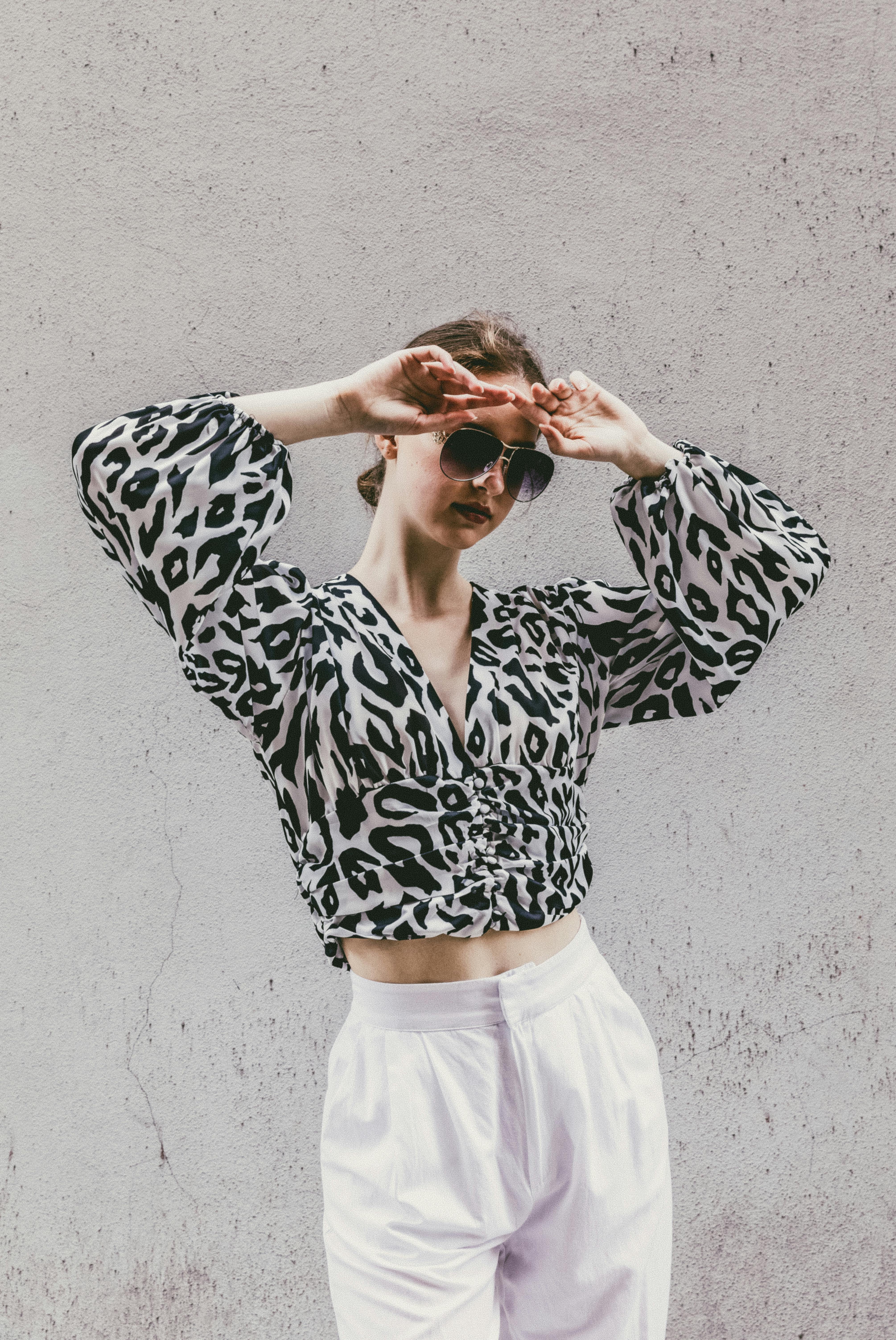 Stylish woman posing in leopard print top and sunglasses against a gray wall.