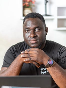 Portrait of a confident black man sitting indoors with hands clasped, wearing a wristwatch.