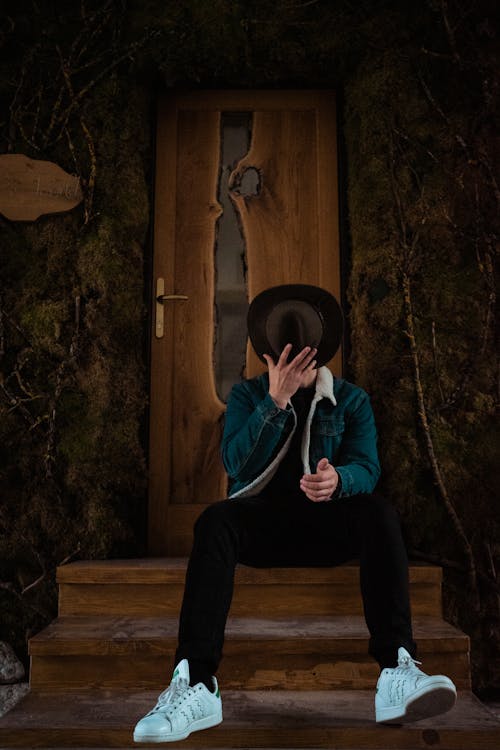 Free Man Taking Off Cowboy Hat While Sitting On Stairs In Front Of Brown Wooden Door Stock Photo