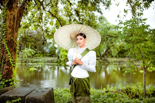 Woman in Sampot and White Lace Blouse with a Green Sash Posing with an Umbrella by the River