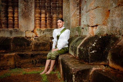 Model in a White Chiffon Embroidered Blouse and a Green Silk Printed Sampot Skirt Sitting in the Ruins of a Historic Temple